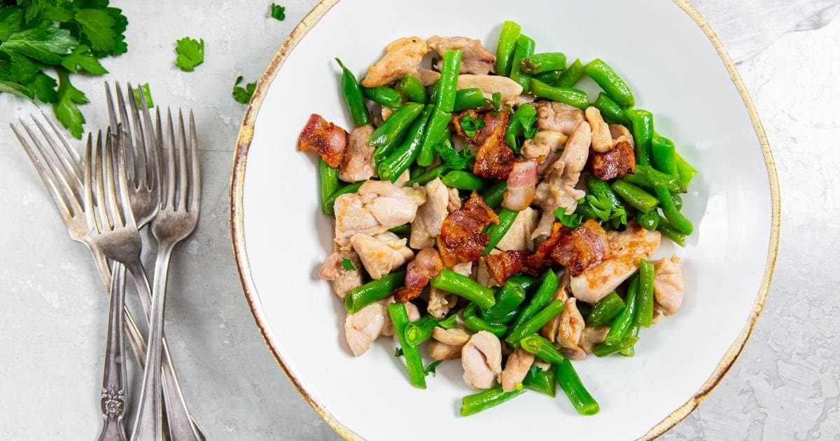 Best Chicken Recipes That Will Have Your Family Begging For More Skillet Chicken Thighs, Bacon, and Green Beans on a Plate Next to a Table Setting of Forks and a Knife