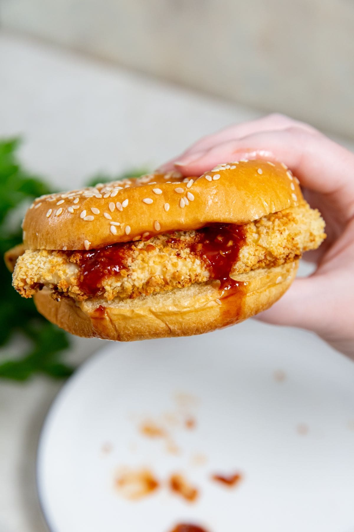 Air Fryer Katsu Chicken Sandwich in a Person's Hand Being Held Above a White Plate with Crumbs and Sauce