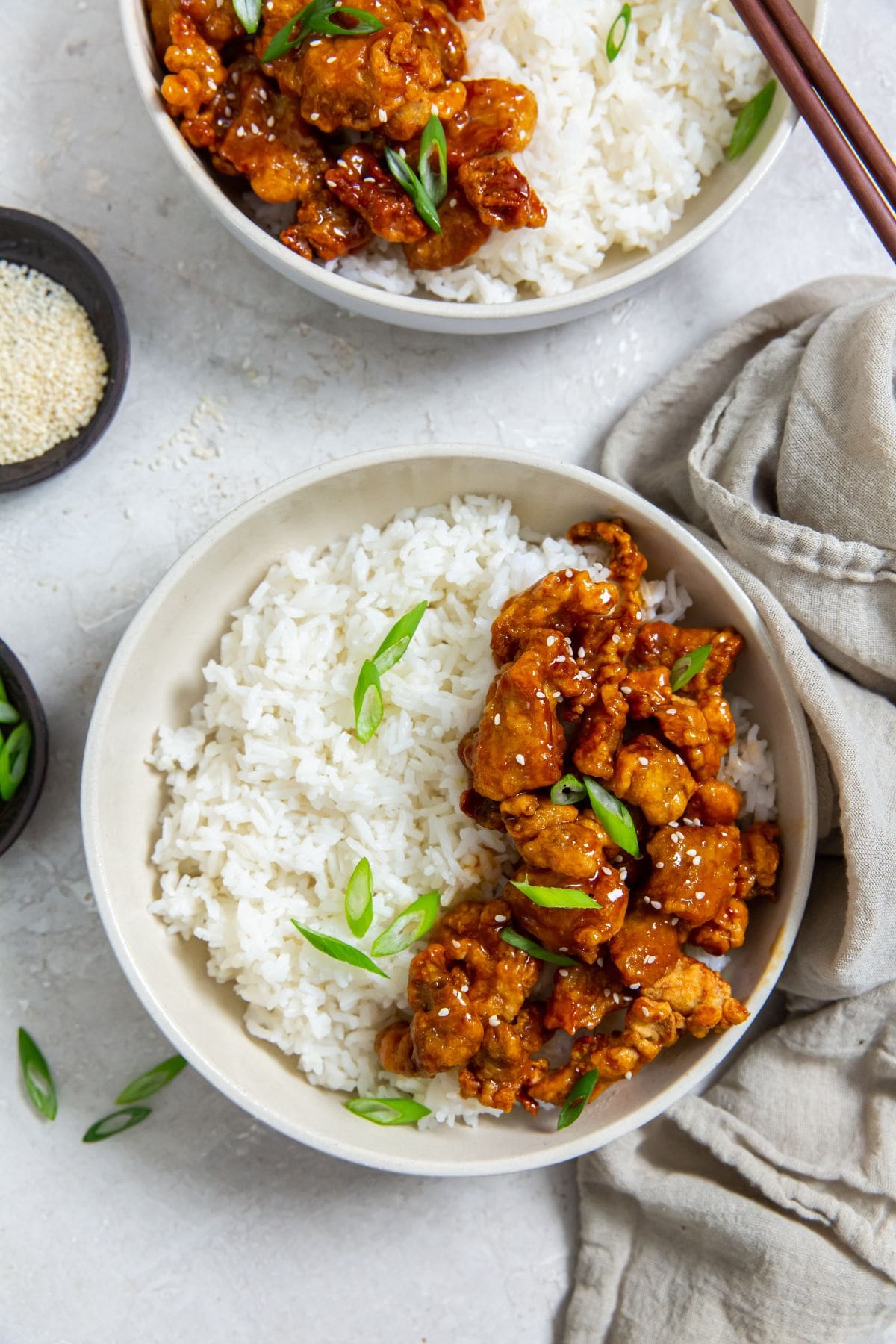 Air Fryer Trader Joe&rsquo;s Orange Chicken white bowl with chicken and rice inside. scallions on top. Gray towel next to it. scallions and sesame seeds in the back. chopsticks on the bowl