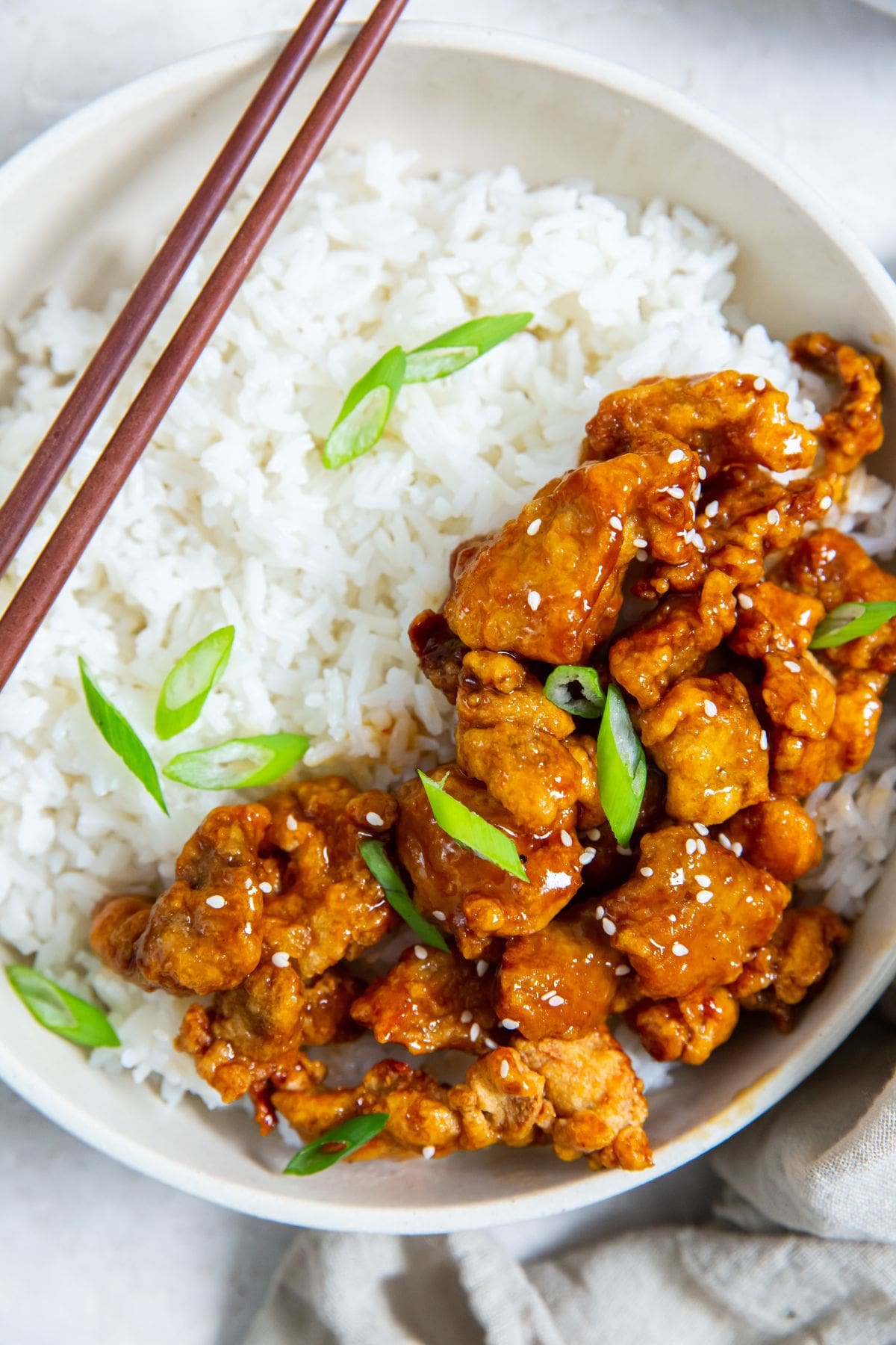 Air Fryer Trader Joe&rsquo;s Orange Chicken white bowl with chicken and rice inside. scallions on top. Gray towel next to it. scallions and sesame seeds in the back. chopsticks on the bowl