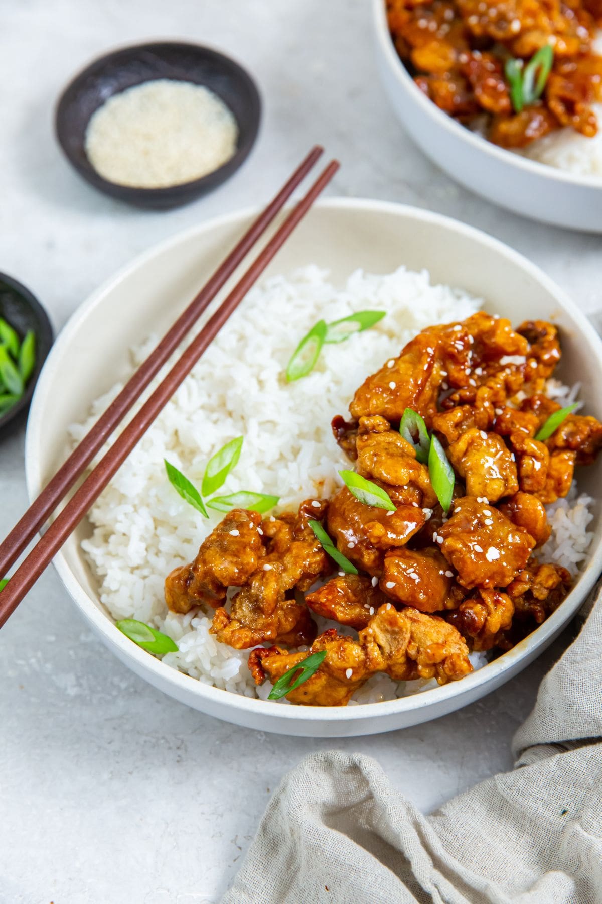 Air Fryer Trader Joe&rsquo;s Orange Chicken white bowl with chicken and rice inside. scallions on top. Gray towel next to it. scallions and sesame seeds in the back. chopsticks on the bowl