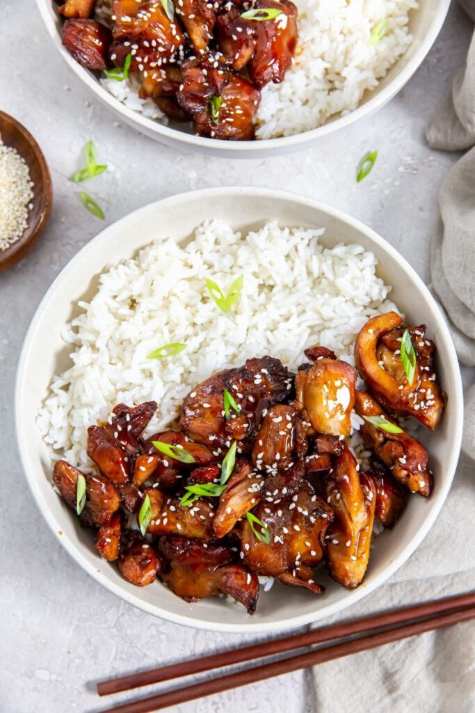 Air Fryer Trader Joe&rsquo;s BBQ Teriyaki Chicken two bowls with chicken and rice inside them. gray towel in the back along with chopsticks and sesame seeds