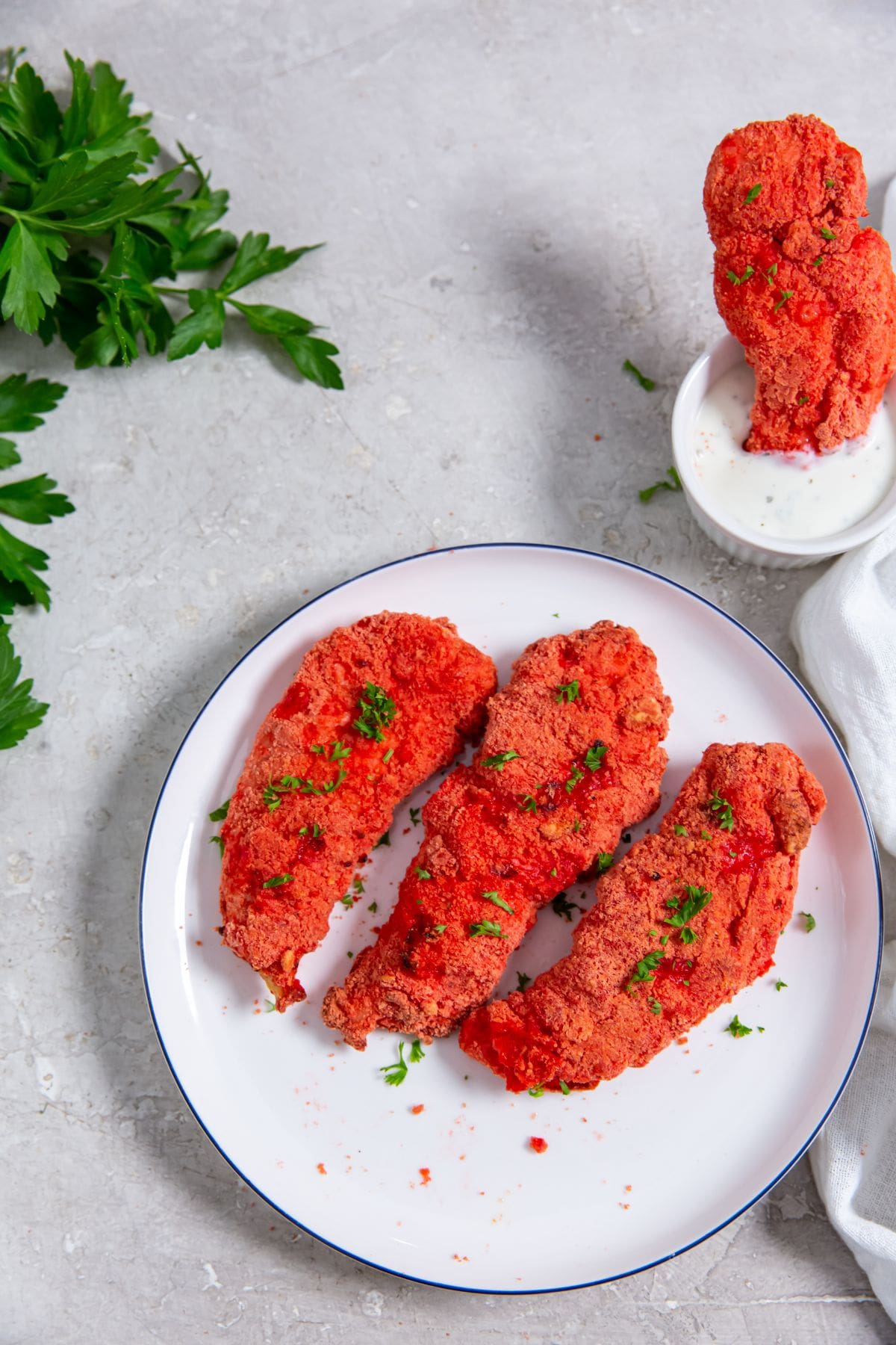 Air Fryer Flamin' Hot Cheetos Chicken Tenders white plate with chicken parsley and white towel next to it. One chicken dunked in ranch