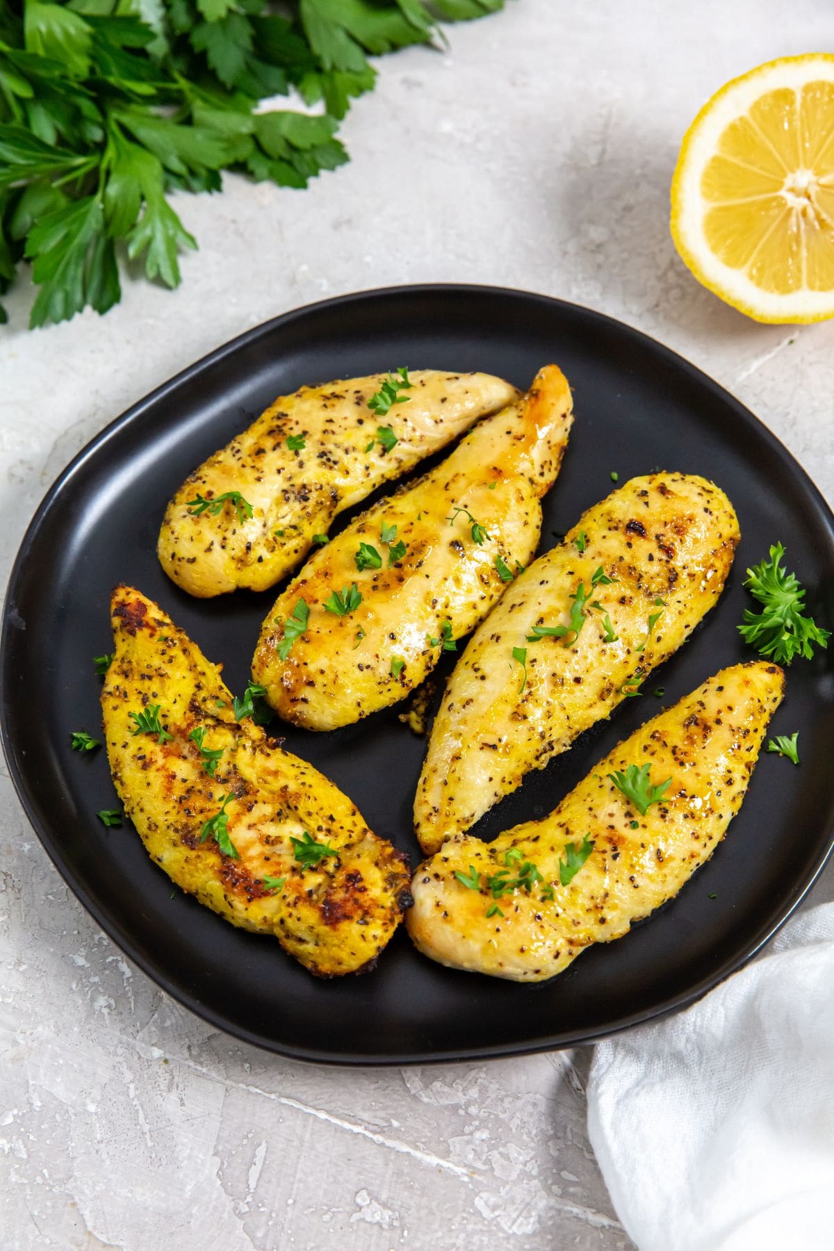 Air Fryer Lemon Pepper Chicken Tenders black plate with chicken on it. parsley, lemon, white towel in the back.