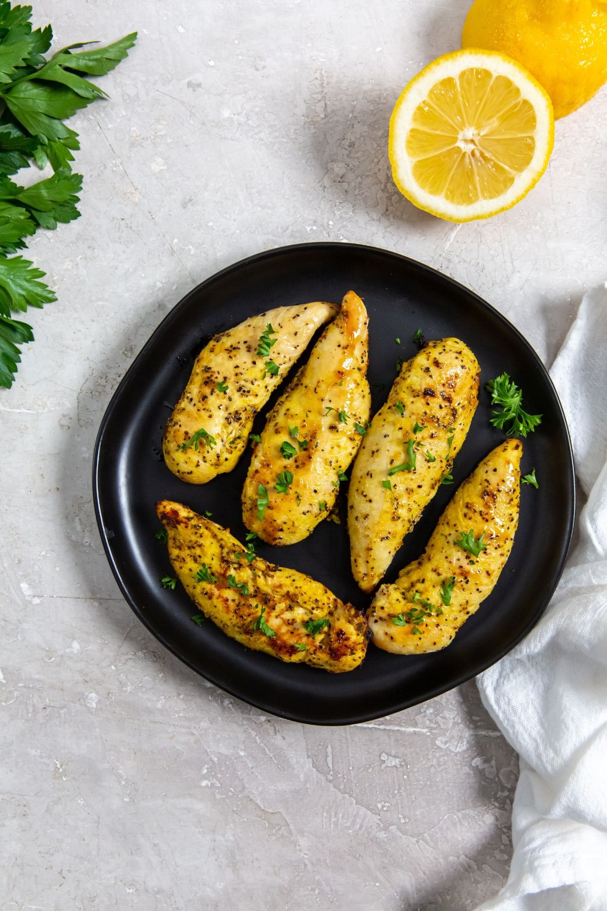 Air Fryer Lemon Pepper Chicken Tenders black plate with chicken on it. parsley, lemon, white towel in the back.