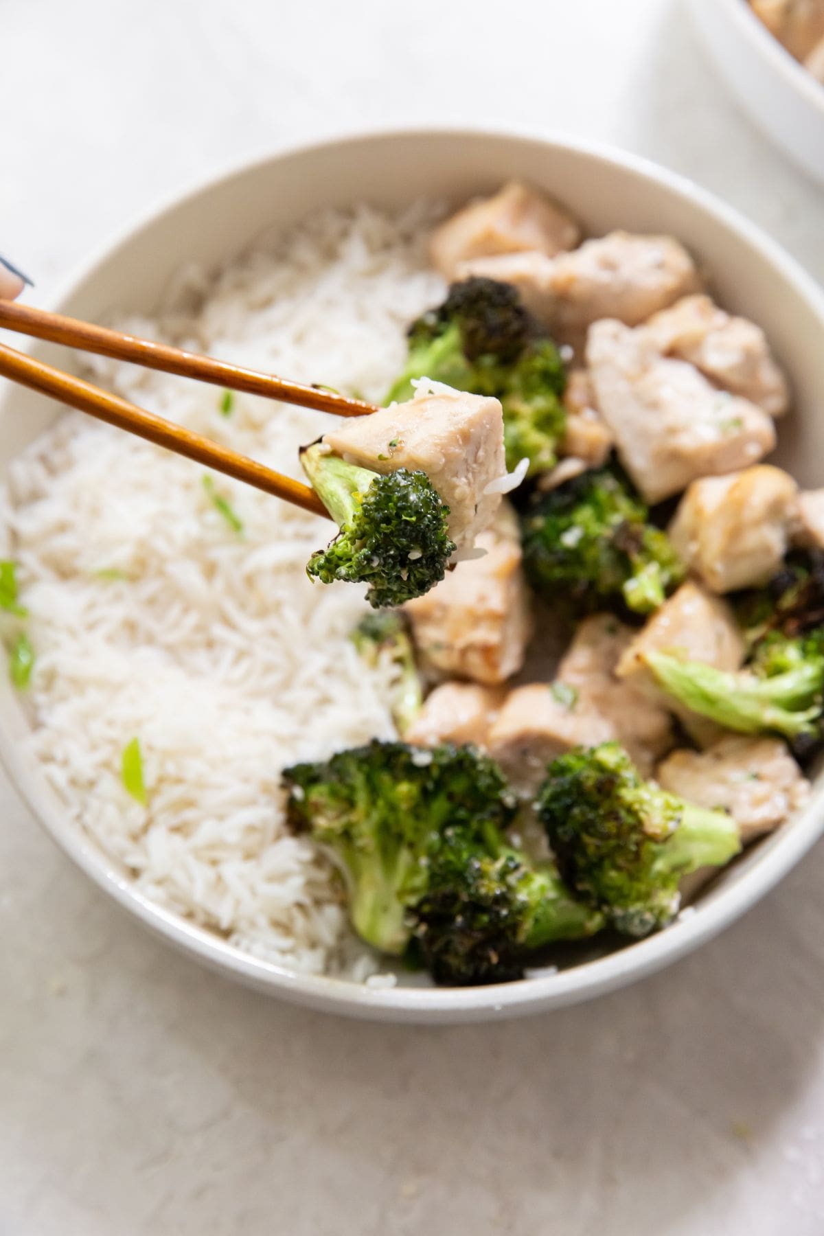 Air Fryer Chicken and Broccoli Chopsticks Holding a Piece of Chicken and a Piece of Broccoli Above a White Bowl with White Rice and More Chicken and Broccoli