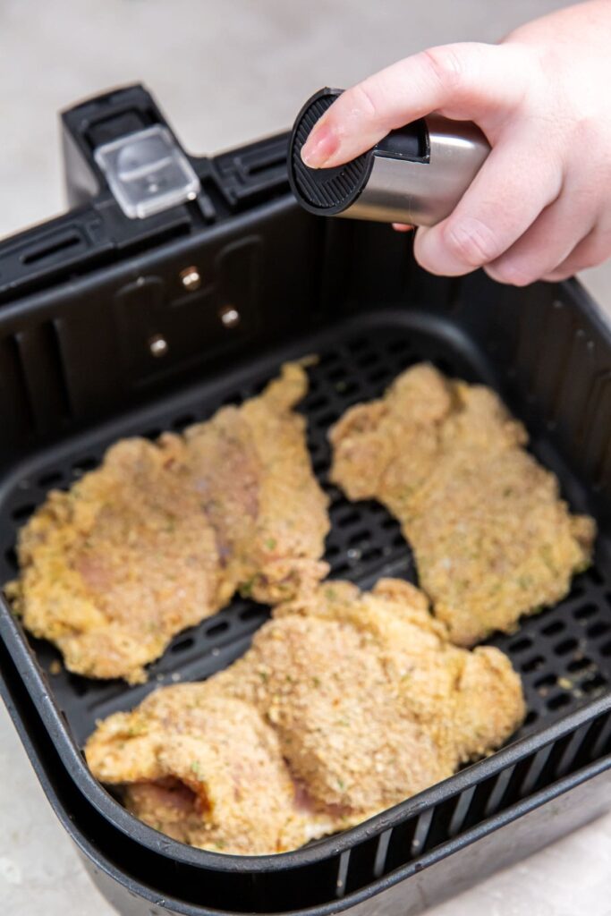 Air Fryer Breaded Chicken Thighs black air fryer with raw chicken inside. getting sprayed with avocado oil
