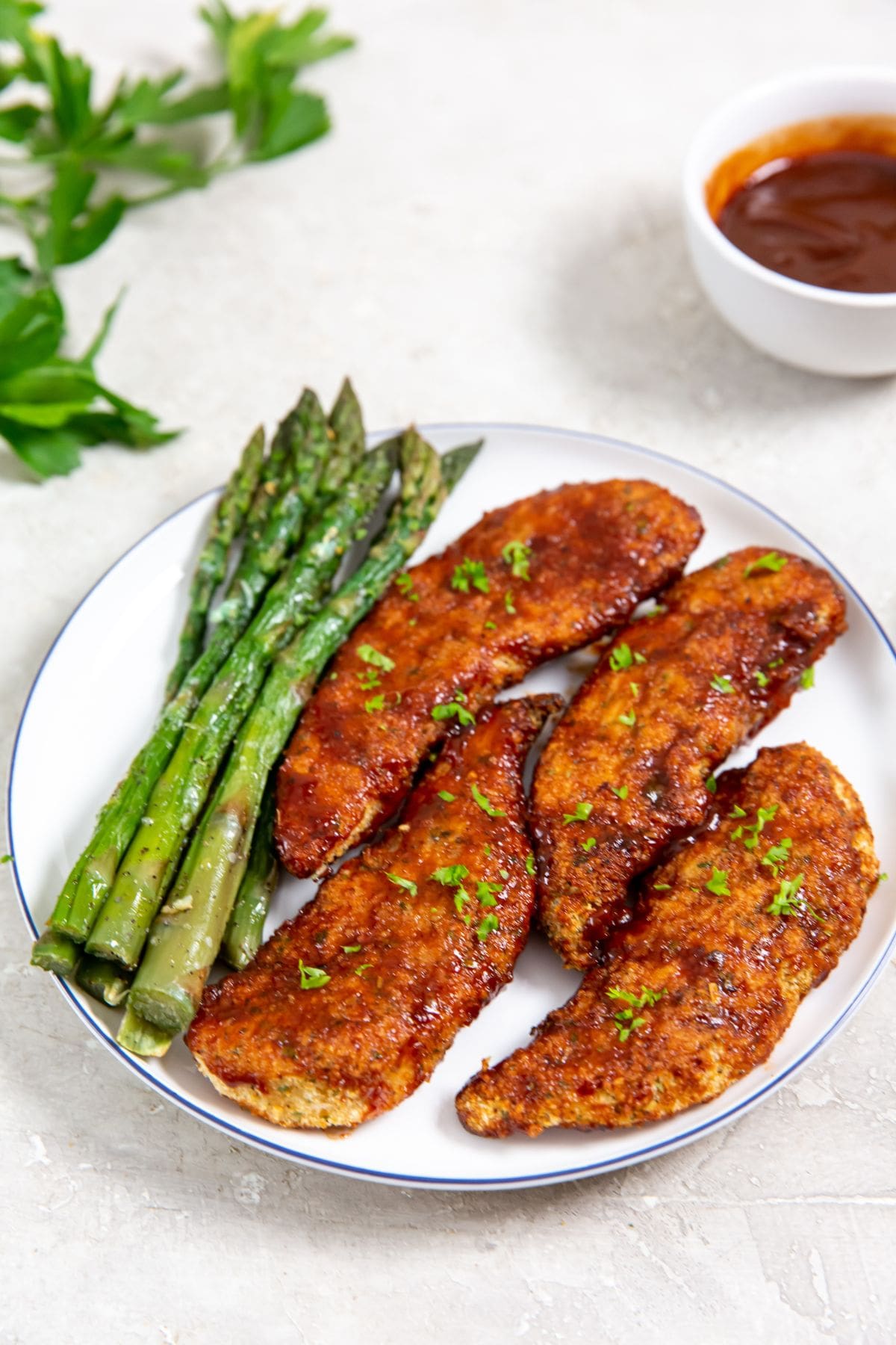 Easy Air Fryer Honey BBQ Chicken Tenders white plate with chicken and asparagus on it parsley and bbq sauce in the back
