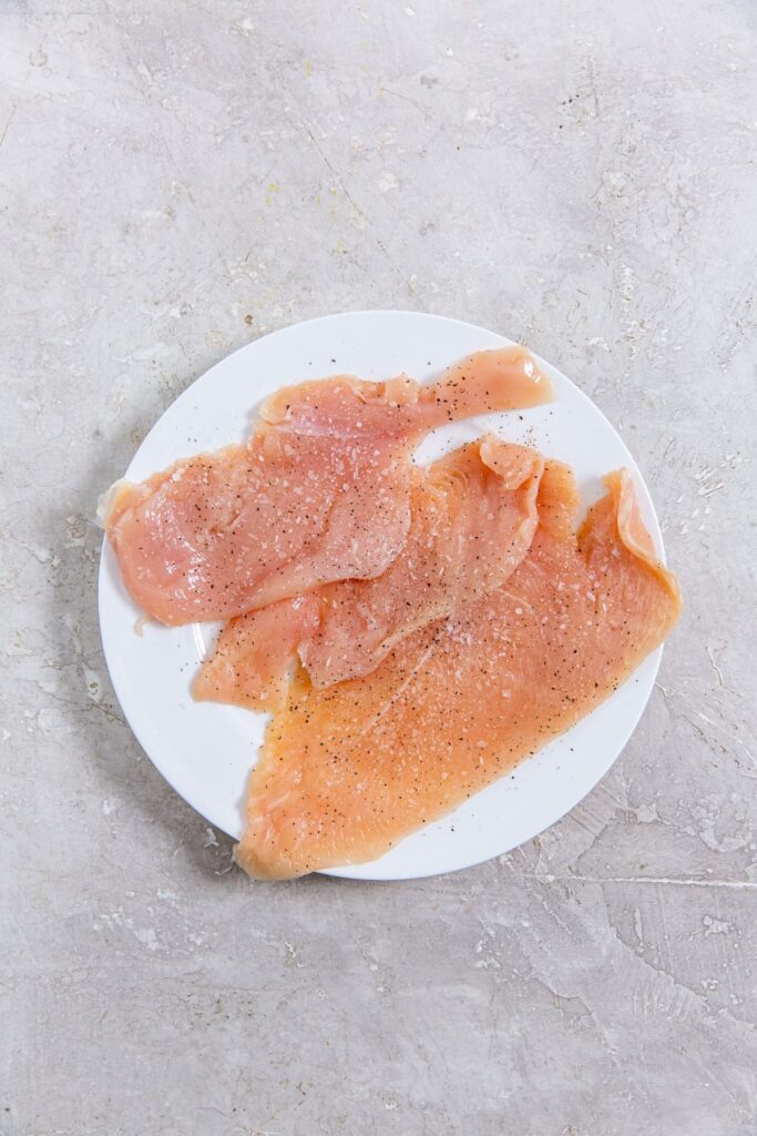 Air Fryer Thin Sliced Chicken Breast raw chicken on a white plate seasoned with salt and pepper.