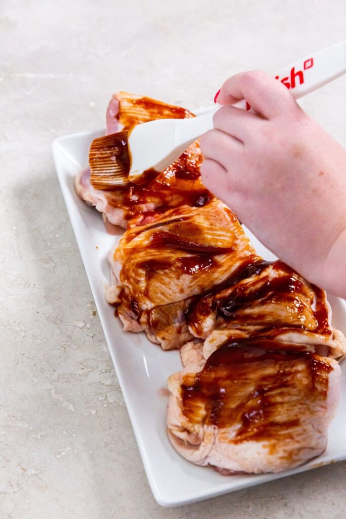 Air Fryer BBQ Chicken Thighs Raw Chicken Thighs on a White Plate Being Brushed with BBQ Sauce