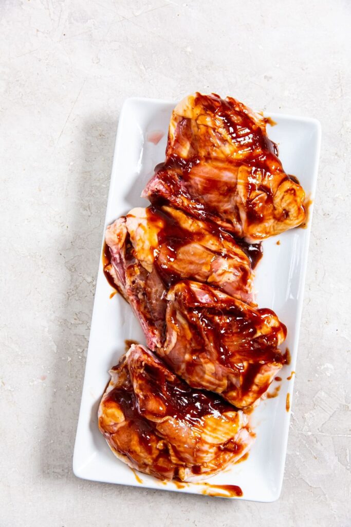 Air Fryer BBQ Chicken Thighs white plate with raw chicken on it. coated in bbq sauce