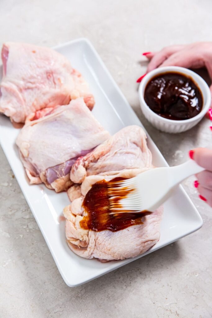 Air Fryer BBQ Chicken Thighs white plate with raw chicken on it. bbq sauce getting brushed on